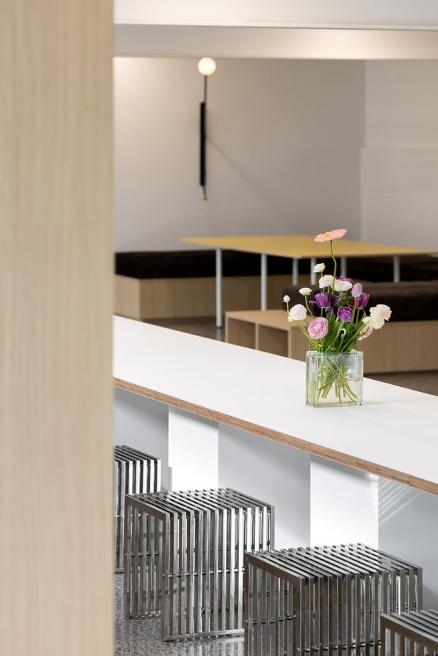 A refined contemporary commercial interior with a split-level or stair-edge seating area. A white counter or balustrade with a timber edge runs through the foreground, overlooking rows of sculptural slatted metal benches or stools on a patterned floor. Behind it are a long timber-toned bench, a yellow tabletop, a minimalist black wall light, and a glass vase of flowers. The space uses restrained neutral finishes, clean lines, and strong geometric repetition.
