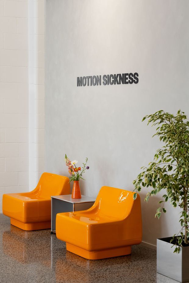 A bright, minimalist waiting or lounge area with two glossy orange sculptural armchairs, a small square side table, a potted tree, and a floral arrangement. The space has a pale grey plaster wall, white painted masonry wall, and polished speckled flooring. A wall sign reads “MOTION SICKNESS.”