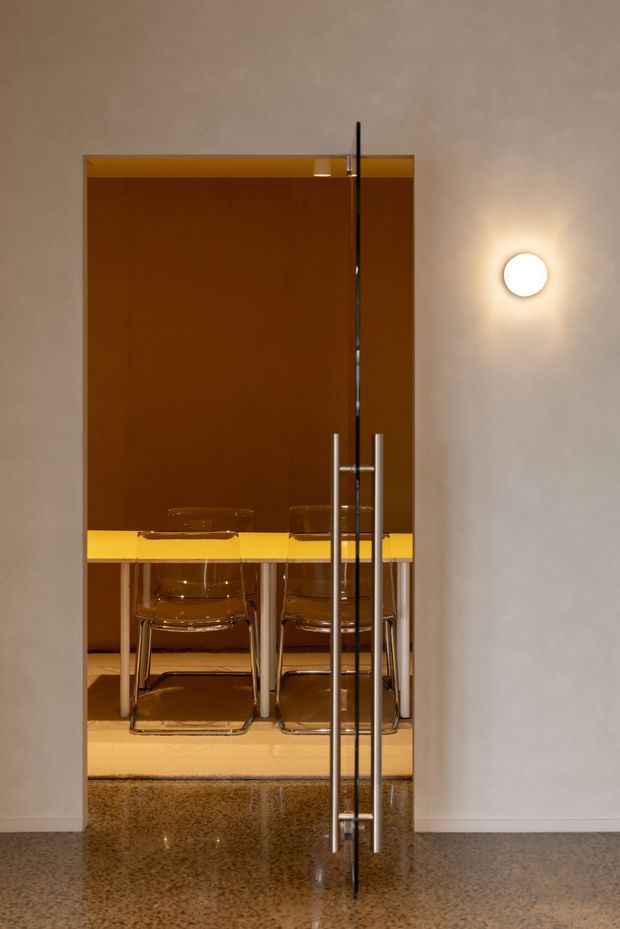 View through a tall glass pivot door into a compact dining room. The room features a glossy amber-brown wall or panel, a bright yellow rectangular dining table, transparent acrylic chairs with chrome frames, polished flooring, and a round wall sconce. The composition has a refined, dramatic, retro-modern appearance with strong reflective surfaces.