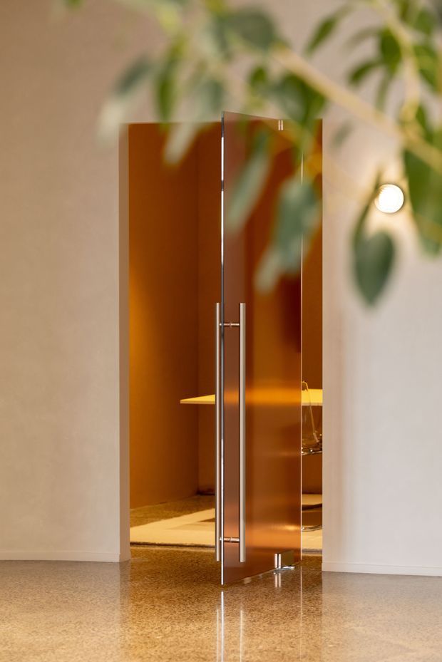 A minimalist interior view of a partially open, full-height pivot door with a reflective warm bronze or amber-toned surface and slim metal handle. The doorway opens into a warmly lit room with a low table or console visible beyond. The surrounding walls are smooth light plaster, and the polished speckled floor reflects the door. Out-of-focus indoor foliage frames the upper foreground, while a small round wall light is visible on the right.