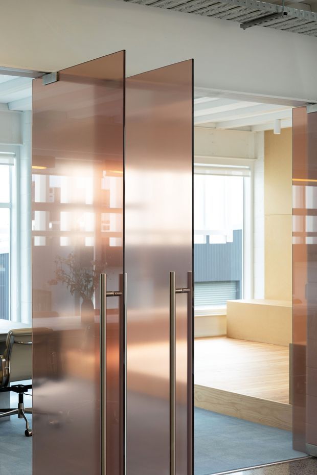 Modern commercial interior featuring tall translucent bronze-tinted glass doors with dark slim frames and vertical metal pull handles. The doors open onto a bright, open-plan workspace with large windows, pale timber flooring, a raised platform or built-in bench, exposed structural ceiling elements, and blue carpet. Office furniture is partially visible at the edges.