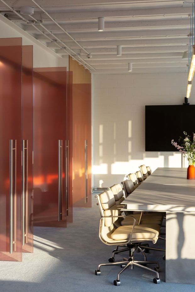 A bright contemporary commercial meeting room with a long light-coloured conference table and upholstered swivel chairs. Tall glossy terracotta-red storage or partition panels with long metal handles line the left side, while a white-painted block wall, exposed white ceiling services, suspended cylindrical lights, and a large black wall-mounted screen define the space. Strong low-angle sunlight creates warm reflections and geometric shadows across the room.