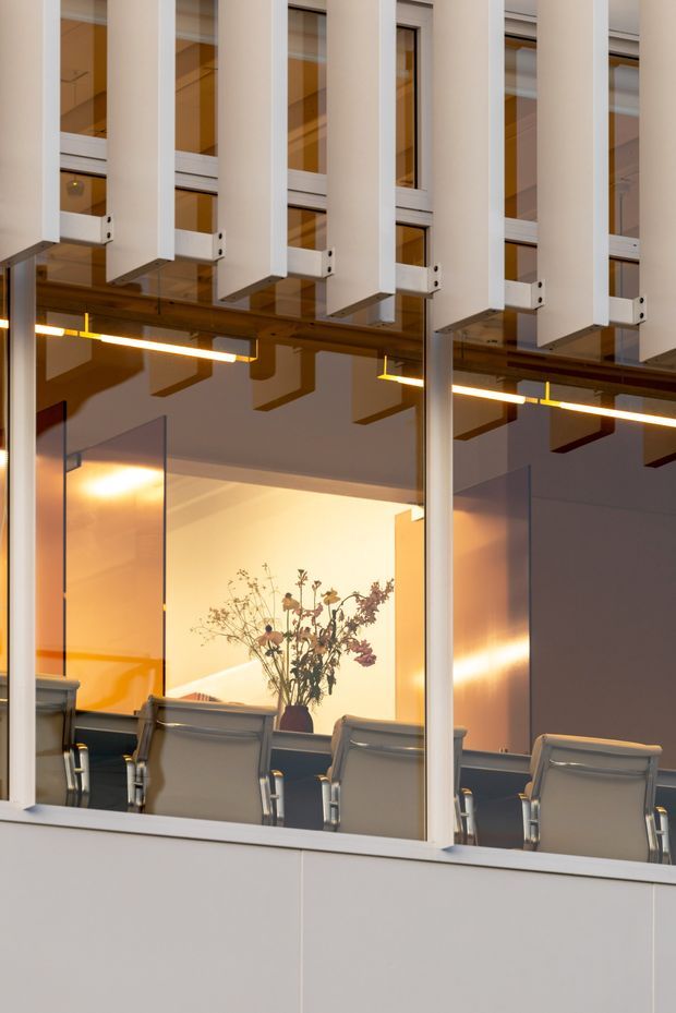 A contemporary commercial interior viewed through a glazed partition, showing a conference or meeting room with a long table, several upholstered office chairs, a vase of dried flowers, warm linear pendant lighting, and large interior glazing. White vertical structural or acoustic elements and exposed ceiling services create a modern office aesthetic.