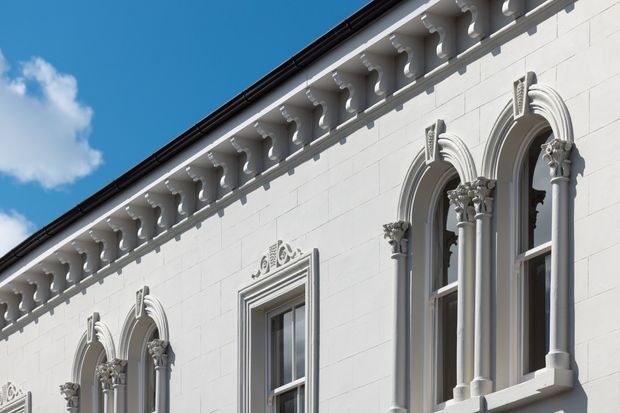 Close-up exterior view of an ornate white masonry facade under a clear blue sky. The building features tall arched windows with elaborate classical mouldings, paired columns with decorative capitals, a richly detailed cornice, and a dark roof edge. The composition highlights historic architectural detailing and symmetry.