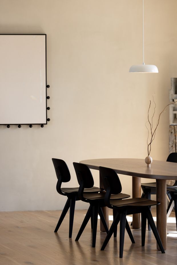 A refined dining area with a long dark timber dining table, sculptural black dining chairs, pale timber flooring, and warm beige walls. A large framed artwork, minimalist white pendant light, and simple vase with branches create a calm, understated composition.