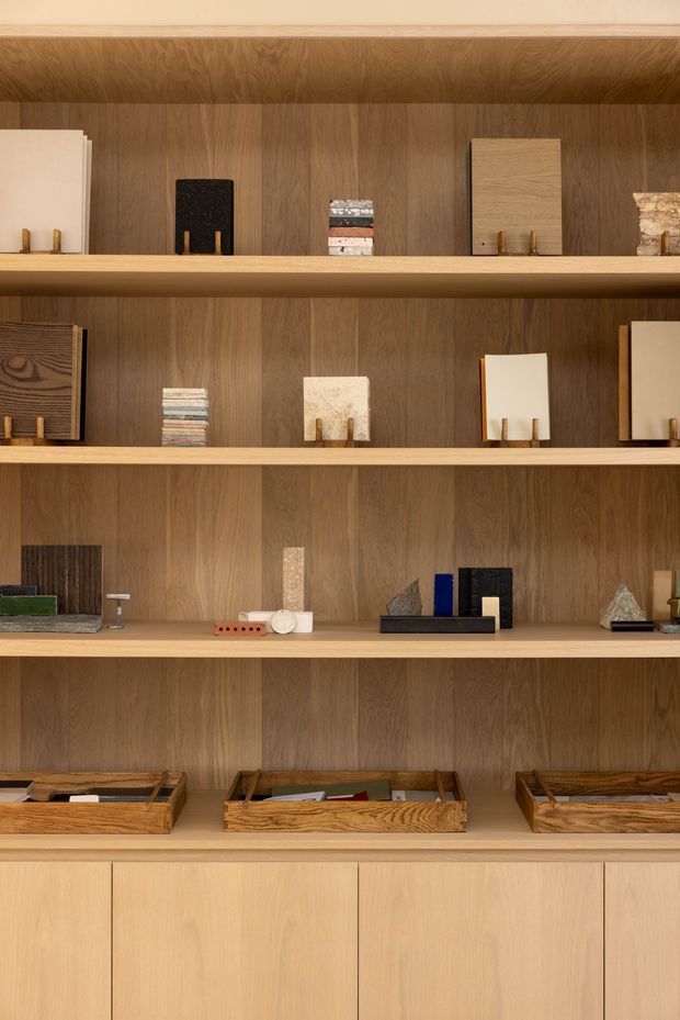 A close-up view of a custom timber display and storage wall with multiple open shelves, vertical wood-grain backing, and lower closed cabinetry. The shelves hold small architectural material samples, books, objects, and trays. The warm natural timber finish creates a refined, curated showroom or workspace display.