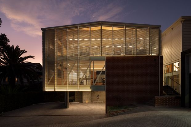 Twilight exterior view of a contemporary multi-level building with a prominent glazed upper volume screened by horizontal louvres. Warm interior lighting reveals an open stair and circulation spaces behind the glazing. A dark brick wall forms the lower facade, with a paved driveway or forecourt and adjacent residential structures visible at the sides.