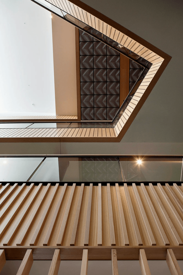 Upward-looking architectural view through a multi-level atrium or stairwell. Warm timber slatted ceiling and wall screening frame a dark geometric patterned feature panel, with black metal-framed glazing and layered balconies creating strong angular lines and a dramatic sense of depth. The image emphasizes fixed architectural detailing rather than furnishings.