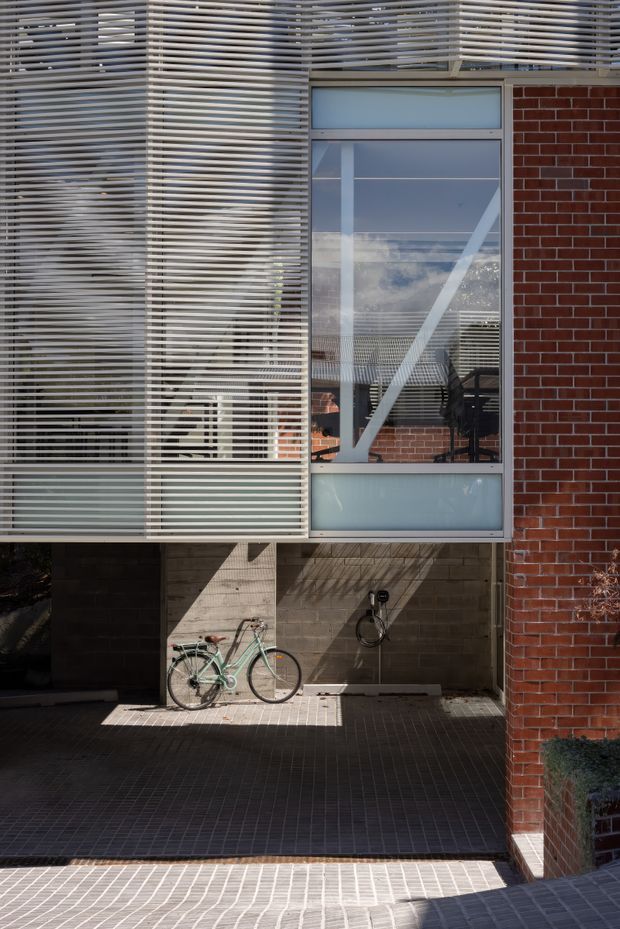 Exterior architectural detail of a contemporary building featuring a prominent horizontal metal louvred screen, large glazed window with visible structural bracing, red brick wall, exposed concrete block, and dark tiled paving. Strong sunlight creates patterned shadows across the facade and ground.