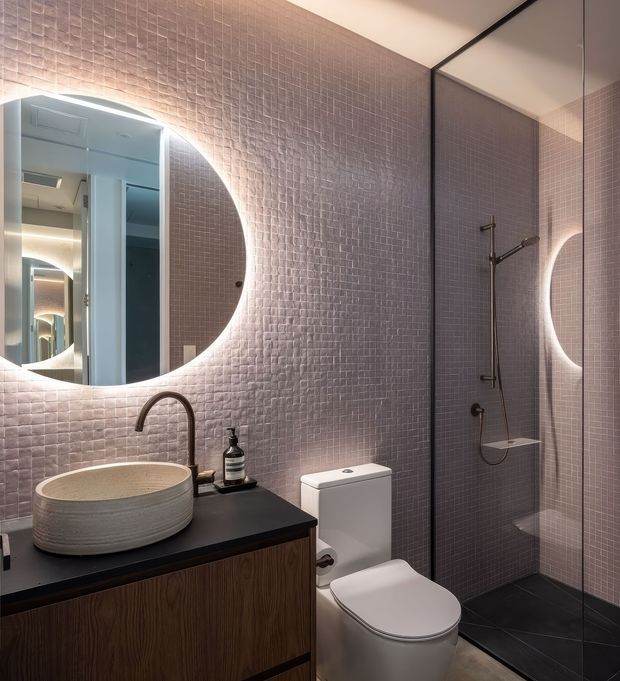 A compact contemporary bathroom with mauve-pink textured mosaic wall tiles, a round backlit mirror, a dark benchtop vanity with a ribbed ceramic vessel basin and dark curved tapware, and a white toilet. A frameless glass shower enclosure occupies the right side, with black floor tiles and warm integrated lighting creating a refined, moody atmosphere.