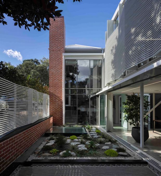 A contemporary multi-level courtyard exterior with a minimalist Japanese-inspired gravel garden, rocks and low planting. The building combines red brick walls, extensive floor-to-ceiling glazing, light-coloured horizontal metal screening and a covered outdoor walkway. A reflective pond or glazed water feature sits near the base of the courtyard, while a large potted tree and surrounding mature trees add greenery. Bright clear daylight emphasizes the crisp modern forms and layered facade.