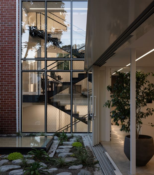 A refined contemporary courtyard interior with a Japanese-inspired dry garden in the foreground, featuring stepping stones, gravel, low planting, and a shallow reflecting pool. A tall black-framed glazed atrium reveals a dramatic open stairway and adjoining building volumes beyond. To the right, a light-filled interior passage with linear recessed lighting, pale flooring, white structural columns, and a large potted tree opens onto the courtyard.