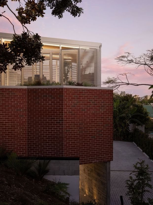 Exterior architectural photograph at dusk showing a contemporary elevated house. A large red brick wall forms the main facade, with a light-coloured upper volume enclosed by horizontal louvres and extensive glazing. The building sits on a sloping landscaped site with a tiled or gridded access surface, while trees frame the view against a pink evening sky.