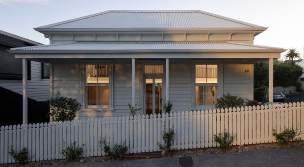 Front exterior view of a traditional single-storey timber-clad residence with a broad hipped corrugated-metal roof, covered front verandah, white picket fence, and vertically proportioned windows and entry door. The muted grey facade and white trim give the home a restrained, heritage-inspired appearance.