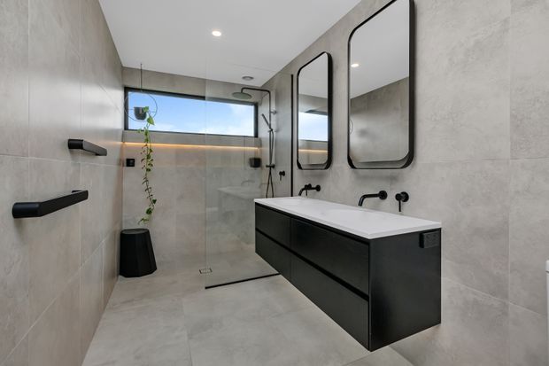 Contemporary bathroom with large-format light grey wall and floor tiles, a floating matte-black double vanity, integrated white basin, black wall-mounted tapware, two rounded-rectangle mirrors, and a spacious open shower. A high horizontal window and subtle trailing plant add natural light and greenery.