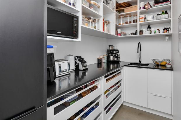 A compact, highly organised walk-in pantry with white open shelving and drawers, a dark stone-look benchtop, integrated microwave, sink, coffee machine, toaster and extensive food storage. The space has a clean contemporary fit-out with neutral finishes and efficient built-in cabinetry.