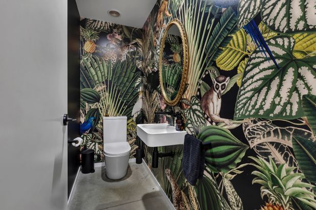 Small, bold powder room featuring a wall-mounted white basin, white toilet, round ornate-framed mirror, black tapware and accessories, and dramatic tropical botanical wallpaper with animal motifs. The room has a grey concrete-look floor and a white ceiling.