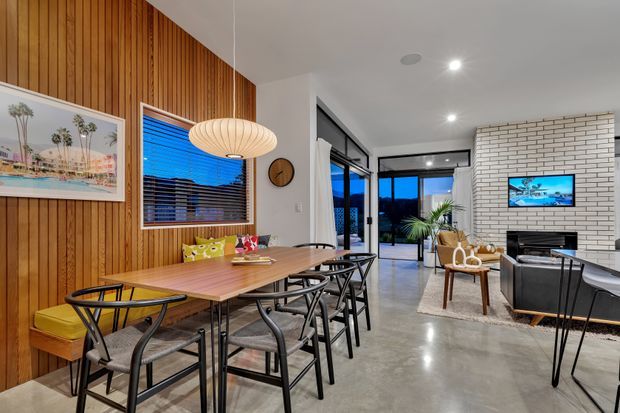 Open-plan dining and living area with a long timber dining table and black chairs, a vertical timber feature wall, sculptural pendant light, polished concrete floor, and a white brick fireplace/media wall. Glazed doors open toward an outdoor area, while contemporary furnishings and artwork add warmth and character.