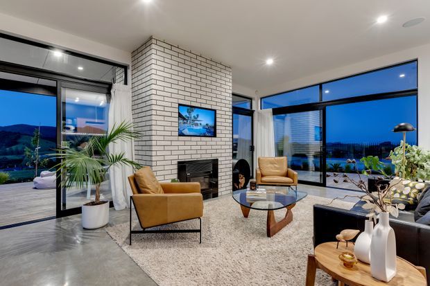 A spacious contemporary living room with polished concrete flooring, a large white-painted brick fireplace wall, floor-to-ceiling black-framed glazing, and views toward a waterfront or coastal landscape at dusk. The room is furnished with caramel leather lounge chairs, a black sofa, a glass coffee table, a textured area rug, indoor palms, and warm decorative lighting.