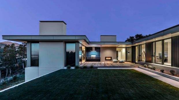 A contemporary single-storey residence arranged around a sheltered central courtyard, photographed at dusk. The home features crisp low-slope rooflines, extensive floor-to-ceiling glazing, dark vertical facade panels, pale rendered walls, and a covered outdoor living area with a fireplace. A lawn occupies the foreground, with a wooded sloping landscape visible beyond the house.