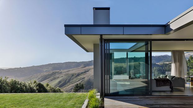 Contemporary flat-roofed house set against a dramatic mountainous landscape. The image focuses on a glazed corner living space opening onto a timber deck, with dark metal framing, expansive sliding glass doors, a neutral sofa, and a projecting roof canopy. The building has crisp rectilinear geometry and strong indoor-outdoor connection.