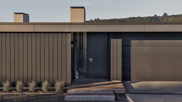 A modern single-storey residential facade with a flat roof, dark charcoal wall cladding, warm vertical timber-toned panels, a recessed glazed entry, and an integrated garage door. Timber steps lead through a minimalist landscaped front garden, with rooftop chimneys and a wooded hillside in the distance.