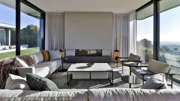 A bright, spacious contemporary living room with floor-to-ceiling corner glazing, sheer curtains, a large sectional sofa, sculptural lounge chairs, and a low rectangular coffee table. The room overlooks a landscaped exterior and distant coastal or hillside scenery. A minimalist fireplace wall with dark built-in joinery anchors the space, while neutral fabrics, dark metal frames, and natural textures create a refined, airy interior.