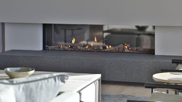 Close-up interior view of a contemporary living room fireplace. A long linear gas fireplace with visible flames is set into a dark textured surround beneath a broad white mantel or wall plane. A reflective glass coffee table and partial seating are visible in the foreground, with a round side table at right. The palette is restrained in white, charcoal grey and light timber, creating a refined modern atmosphere.