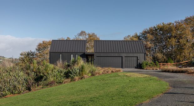 Exterior view of a dark charcoal-clad contemporary house composed of two simple pitched-roof volumes, with a large integrated garage door and smaller vertical windows. The home is set within a landscaped suburban or rural site with native-style planting, lawn, gravel driveway, mature trees, and a timber fence under a clear blue sky.