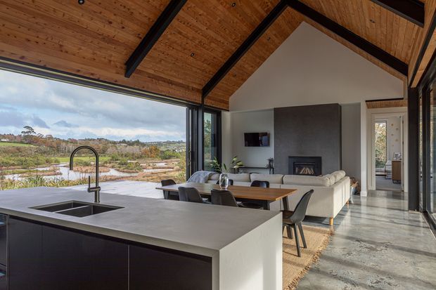 Open-plan contemporary kitchen, dining and living space with a large island and integrated sink in the foreground. The room features a dramatic pitched timber-lined ceiling with exposed dark structural beams, full-height glazing opening to a natural landscape, polished concrete flooring, dark kitchen cabinetry, and a living area with a fireplace and pale sectional sofa.