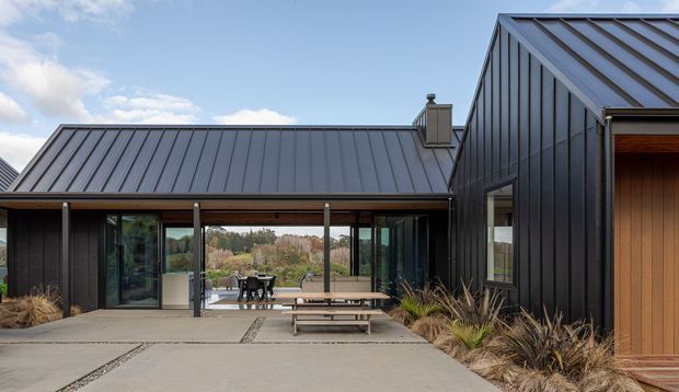 Exterior view of a contemporary, single-storey residence with interconnected steeply pitched and gabled roof forms. The dark vertical-clad facade is paired with standing-seam metal roofing, extensive glazed openings, and a timber-clad entry door. A concrete terrace with outdoor dining furniture fronts landscaped garden beds, with a semi-rural treed outlook beyond.