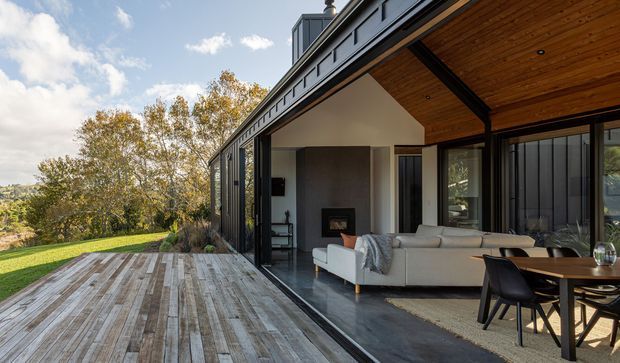 A contemporary indoor-outdoor living and dining space opening onto a large weathered timber deck. The pavilion has extensive black-framed glazing, a dramatic sloping timber-lined ceiling and exposed dark structural beams. Inside, a light sectional sofa faces a fireplace, with a dark dining table and chairs beside the glazing. The setting overlooks a landscaped, tree-filled rural or suburban site.