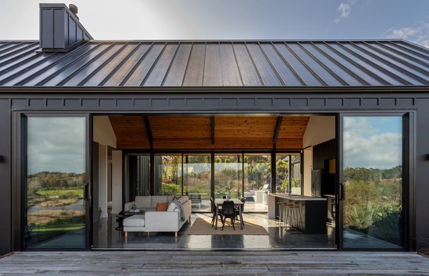 Exterior view of a contemporary single-storey home with a dark standing-seam metal roof and dark framed glazing. Large open sliding doors reveal a combined living, dining and kitchen interior with a timber-lined ceiling, light furnishings, and extensive views toward a green rural landscape. A timber deck spans the foreground.