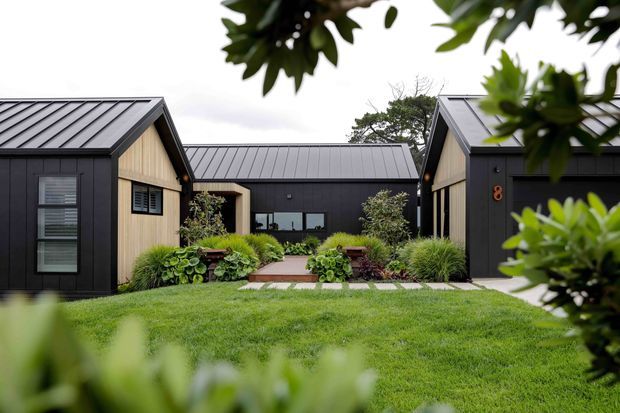 Exterior view of a contemporary, low-rise compound arranged around a landscaped courtyard. Several dark charcoal-clad gabled buildings feature standing-seam metal roofs, warm timber accent walls, large glazing, and a central planted entry path. The foreground is a manicured lawn with dense ornamental planting and leafy vegetation framing the view.