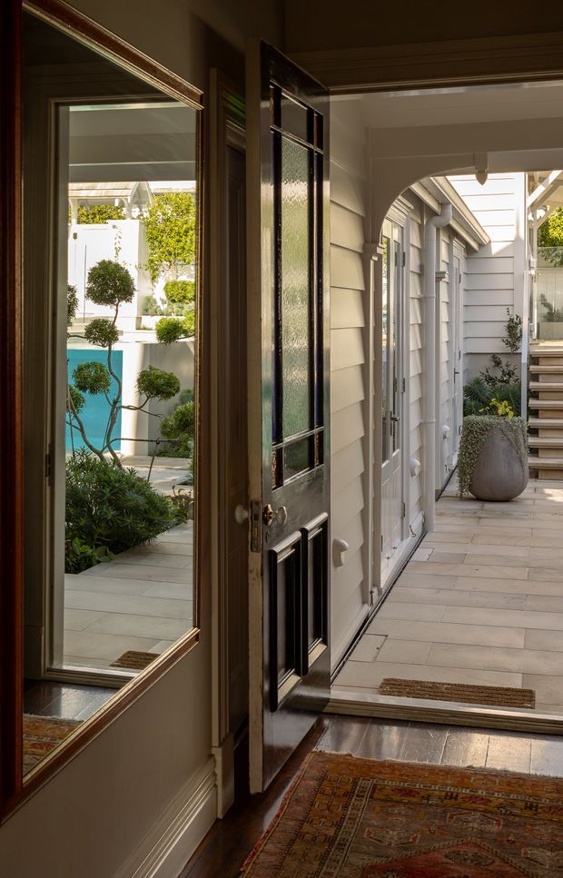 A refined residential entryway opening onto a covered courtyard walkway. The scene features a dark glazed timber front door, traditional trim and wall panelling, a large wall mirror, patterned runner rugs, pale horizontal weatherboard cladding, and a tiled exterior passage leading toward steps and landscaped poolside planting. The architecture combines classic detailing with a light contemporary courtyard setting.