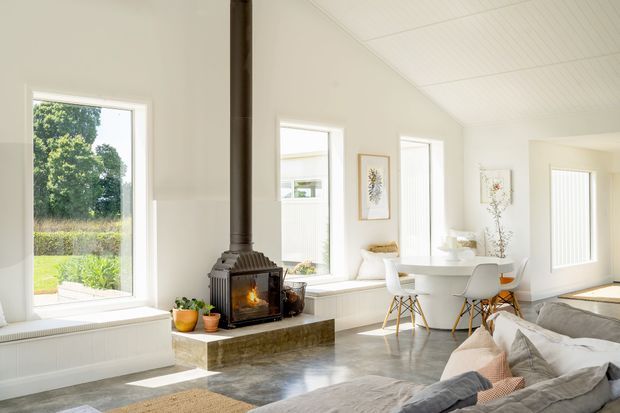 Bright open-plan living and dining space with white walls, a white timber-lined ceiling, broad windows, built-in window seating, and a freestanding wood-burning stove on a stone hearth. A white dining table and modern chairs sit beside a neutral sectional sofa, creating an airy contemporary interior with warm natural light.