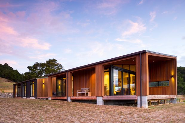 A low, elongated modern timber-clad building set in a rural landscape at dusk. The facade combines deep reddish-brown vertical timber boards, extensive dark-framed glazing, recessed covered verandas, and projecting roof planes. Warm interior lighting glows through the windows, with a small outdoor seating area on the timber deck. The structure sits slightly elevated on concrete supports, with wooded hills and open dry grassland surrounding it.