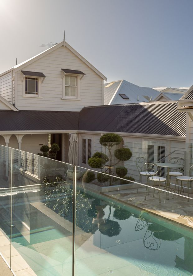 A sunlit courtyard pool area viewed from an elevated terrace, featuring a glass pool safety barrier, pale tiled paving, sculpted topiary, ornate outdoor café furniture, and a surrounding white weatherboard house with pitched roofs, dormer-like windows, verandah details, and dark metal roofing.