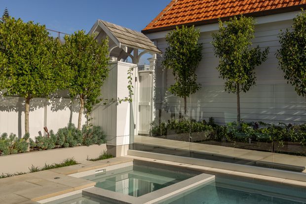 A refined courtyard pool area beside a white weatherboard-clad building with a terracotta tiled pitched roof. The scene includes a compact swimming pool with pale stone paving, raised tiled edges, a glass safety balustrade, manicured espaliered trees, white fencing, and a small gabled garden structure.
