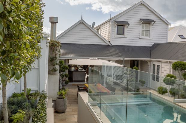 A landscaped courtyard viewed from an elevated position, featuring a compact swimming pool enclosed by frameless glass balustrades, a covered outdoor entertaining area, and a white weatherboard multi-level house with gabled roof forms and dark corrugated roofing. Potted plants, clipped topiary, paving, and outdoor seating create a refined residential setting.