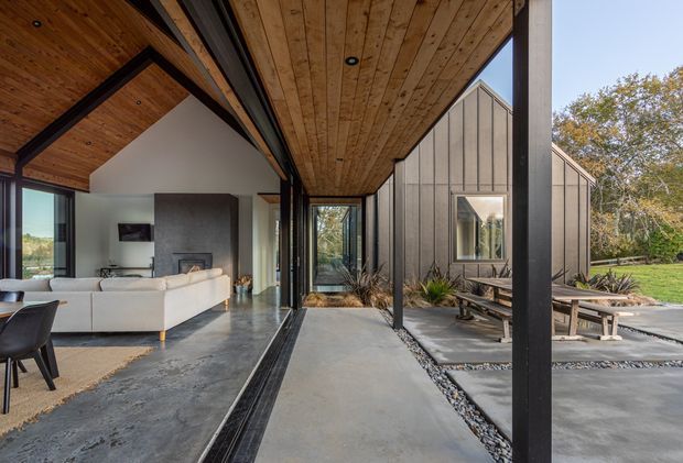 Contemporary indoor-outdoor living space with expansive sliding glass doors opening from a living room onto a covered concrete terrace and landscaped courtyard. The interior has a white sectional sofa, dark feature fireplace wall, polished concrete flooring, and a warm timber-lined ceiling supported by dark steel or timber beams. The exterior wing features vertical grey cladding, concrete paving, gravel drainage strips, and a picnic-style timber table, surrounded by mature planting.