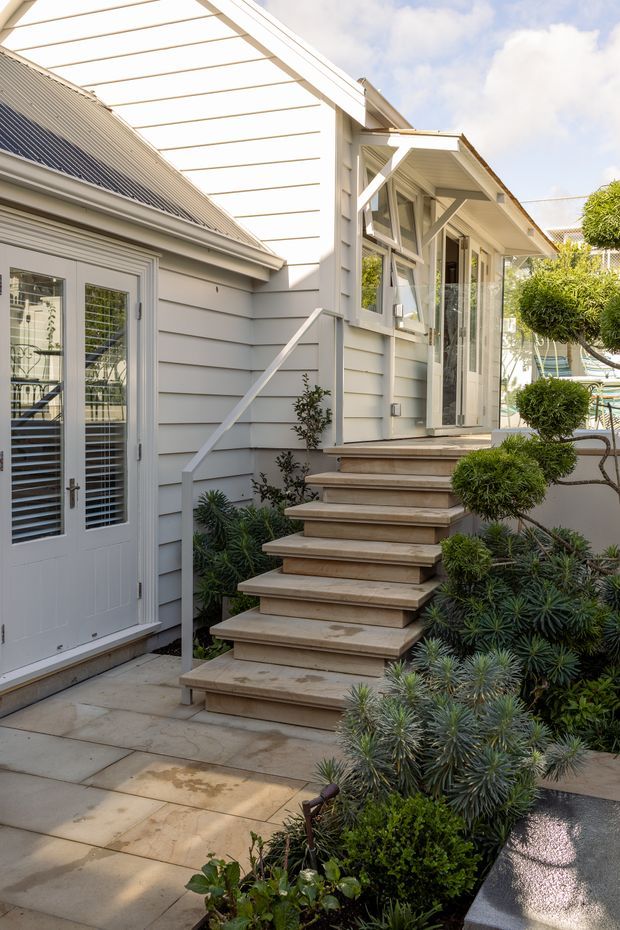 A bright exterior view of a compact landscaped courtyard or garden passage beside a light-grey weatherboard house. Broad timber steps with a simple white handrail lead to a glazed entry or sunroom, while white-framed glazed doors and windows, a corrugated metal pitched roof, and a translucent covered roof structure are visible. Carefully clipped shrubs and layered planting surround pale paved surfaces.