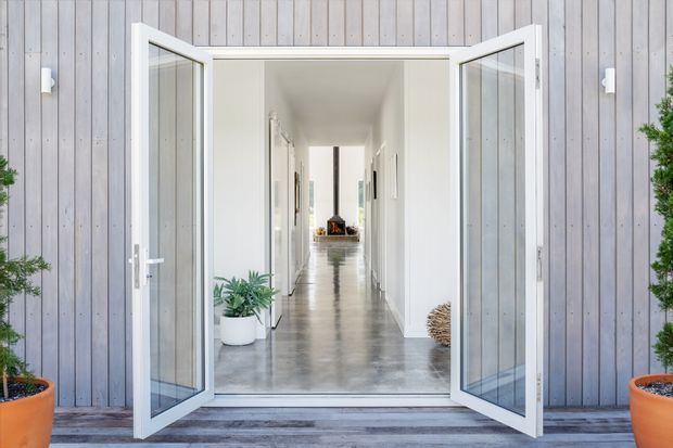 A bright modern entry viewed from outside through wide-open white-framed glazed double doors. The entrance opens into a long minimalist hallway with white walls, polished grey concrete flooring, simple artwork, indoor plants, and a distant living area. The exterior facade is clad in vertically arranged weathered grey timber boards, with timber decking and potted greenery framing the doorway.
