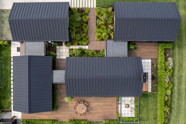 Aerial top-down view of a contemporary residential property composed of several dark metal-clad roof volumes arranged around landscaped garden courtyards. Timber decking, paved paths, planted borders, outdoor dining furniture, and a central garden walkway are clearly visible. The image focuses on the building roof forms and integrated outdoor spaces.