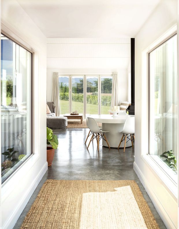 Bright, airy interior view through a glazed entry passage into an open-plan dining and living area. White walls and ceiling, extensive floor-to-ceiling windows, polished concrete flooring, a white dining table with sculptural white chairs, a neutral sofa, indoor plants, and a natural woven runner create a clean contemporary aesthetic. A black vertical flue or structural element provides contrast, while landscaped grounds are visible through the glazing.