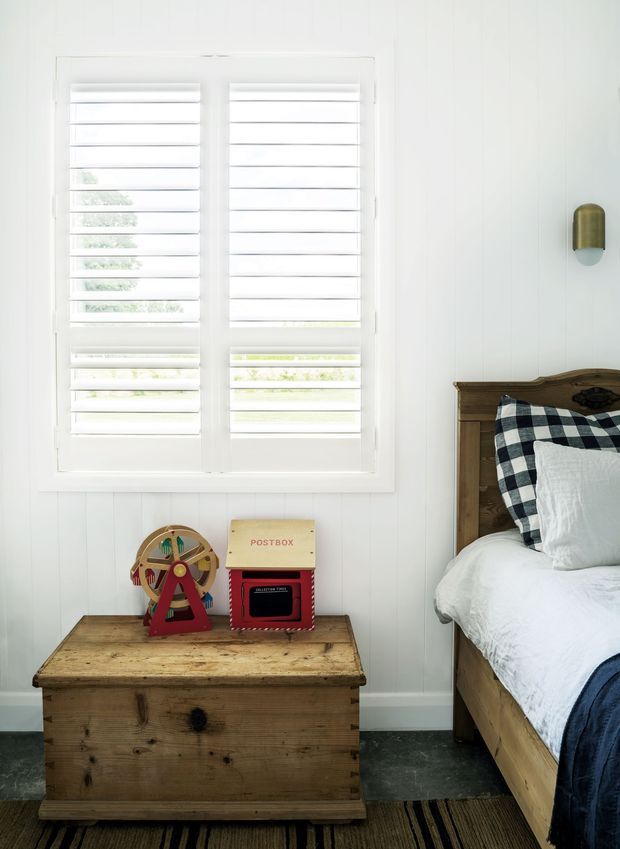 Bright, compact bedroom with white vertical wall panelling and plantation shutters over a large window. A rustic wooden blanket chest sits beneath the window with children's toys on top, while a dark timber bed with white linen, a navy throw, and a checked cushion occupies the right side. A small brass wall sconce adds a warm accent.