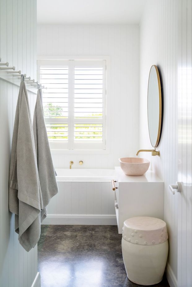 Bright, compact bathroom with white vertical wall panelling, a freestanding bathtub beneath plantation shutters, a white vanity with a round vessel basin, brushed brass tapware, an oval wall mirror, and a small upholstered stool. The dark polished floor provides contrast against the predominantly white and soft neutral palette.