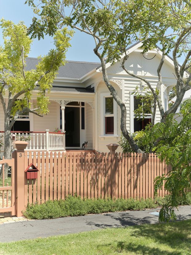 Street-facing view of a detached heritage-style house with a pale painted weatherboard facade, decorative fretwork and ornate porch detailing, a corrugated metal gabled roof, front veranda, timber picket fence, and mature garden planting. The home has a warm peach-pink fence and cream exterior, with a footpath and lawn in the foreground.