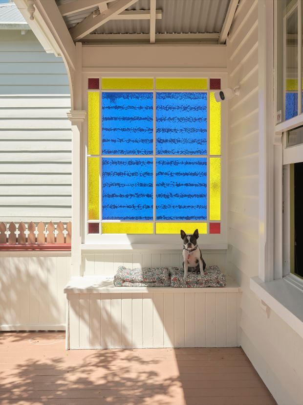 A bright enclosed veranda or sunroom with pale painted weatherboard walls, a corrugated roof with exposed timber framing, and a built-in window seat beneath a striking stained-glass window in blue, yellow, and red. White decorative trims, timber decking, and adjacent exterior glazing reinforce the traditional character. A small dog sits on the cushioned bench.