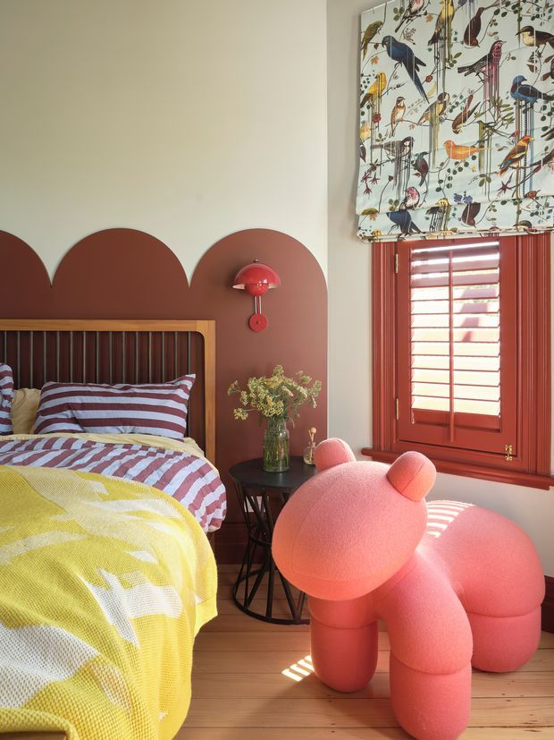 A colourful, playful bedroom featuring a timber bed with striped bedding, a terracotta scalloped painted feature wall, matching red-toned timber window and shutters, and a patterned botanical-and-bird roller blind. A large pink sculptural toy-like chair or seating piece dominates the foreground, with a small black side table and vase beside the bed. Timber flooring and strong sunlight create a warm, cheerful interior.